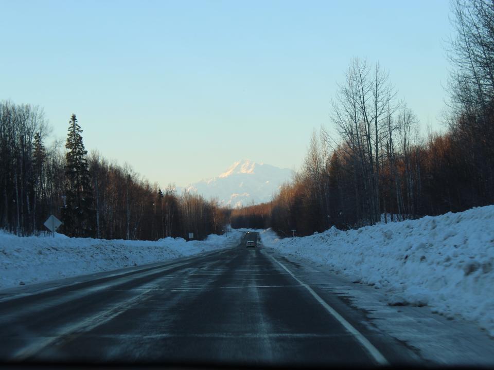 A clear road in Alaska, lined with trees and with mountains in the background