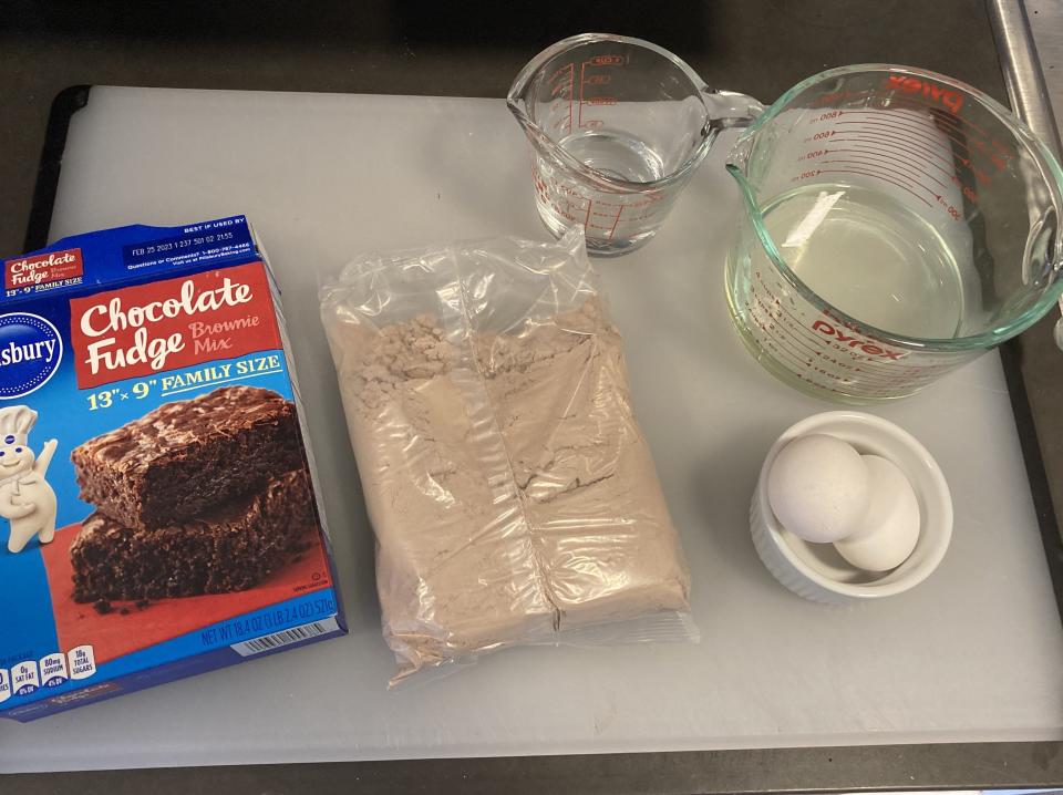 Pillsbury Chocolate Fudge brownie mix box, brownie mix in a bag, a measuring cup of oil, a measuring cup of water, and a small bowl with two eggs on a cutting board
