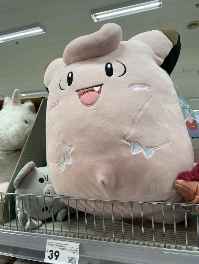 Large plush Clefairy toy sits on a store shelf, priced at $39. Nearby is a smaller gray cat plush