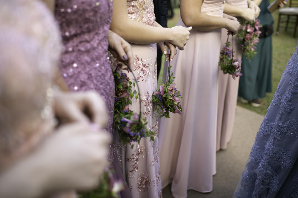 Bridesmaids hold small bouquets of flowers in a wedding procession and wear elegant dresses in various styles with elaborate details and embellishments.