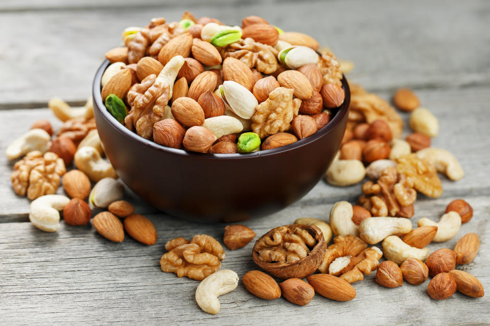 A bowl filled with assorted nuts, including almonds, cashews, pistachios, and walnuts, placed on a wooden surface with more nuts scattered around