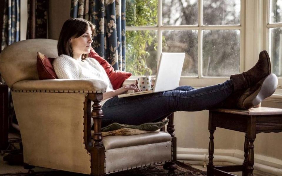 A young woman relaxing at home with her feet up using a laptop - Mint Images Limited / Alamy