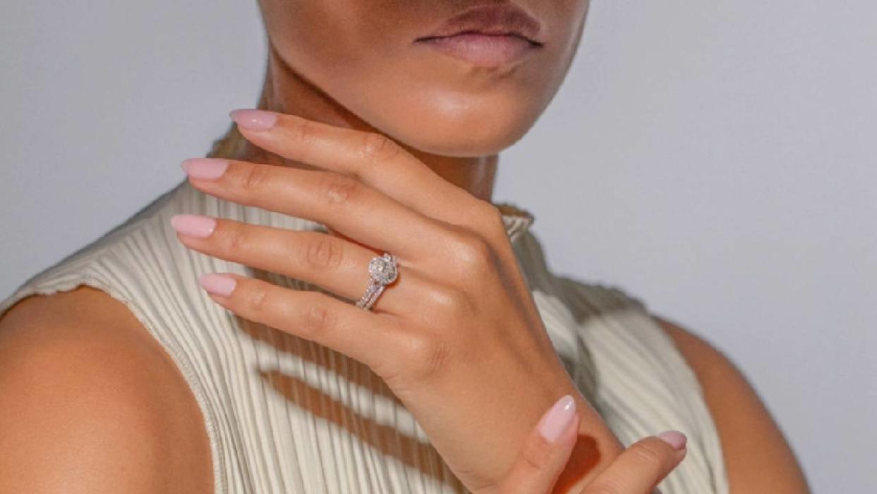  A close up of a woman's hand close to her face, wearing a large, diamond engagement ring from Ernest Jones. 