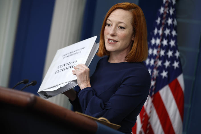 Jen Psaki stands at the briefing room podium, with the Stars and Stripes in the background, holding up a white binder marked COVID-19 Funding..