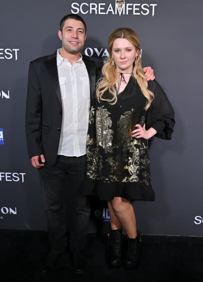 A man with short brown hair in a suit posing with his arm around a woman with long blond hair in a dress