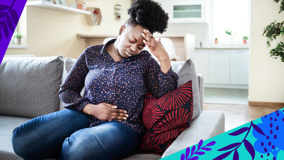 A woman sits uncomfortably on a couch, with one hand pressed to her forehead and the other rubbing her stomach.