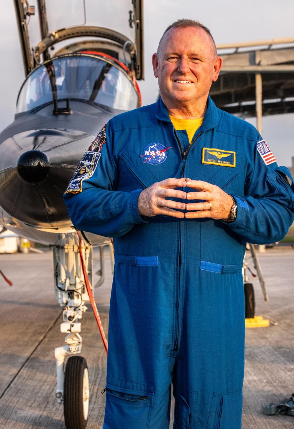an astronaut in flight suit touching his hands in front of his chest. behind him is a jet with cockpit open