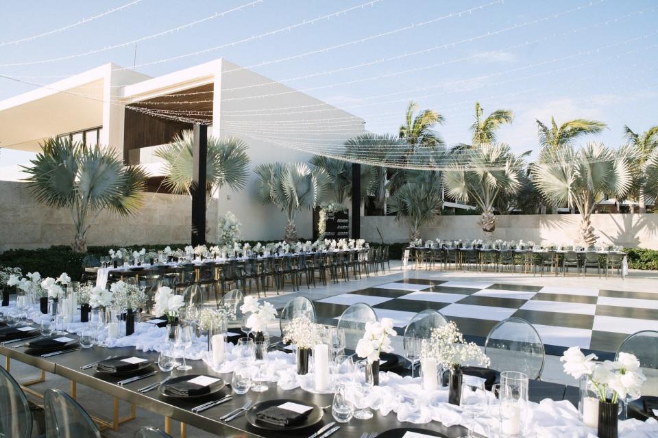 Long tables surround a black-and-white checkerboard floor with lights hanging atop it at an outdoor wedding ceremony.