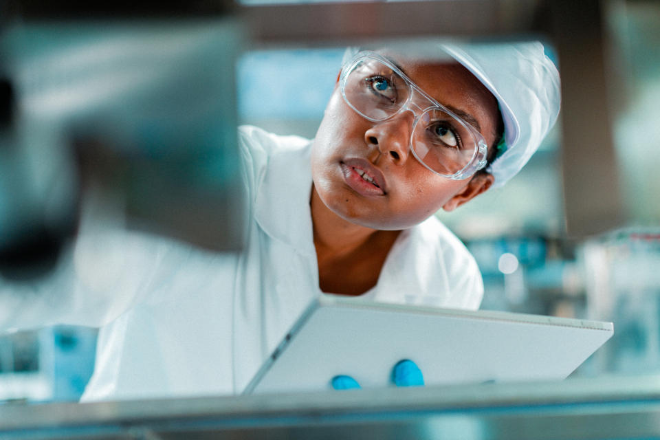 A person in safety goggles, gloves, and a lab coat inspects machinery in a laboratory or industrial setting, focusing intently on the task