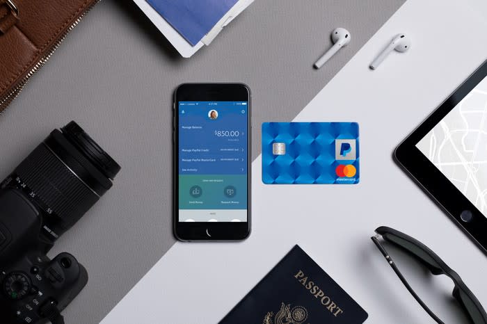 The PayPal app on a smartphone on a desk with a PayPal-branded Mastercard nearby.