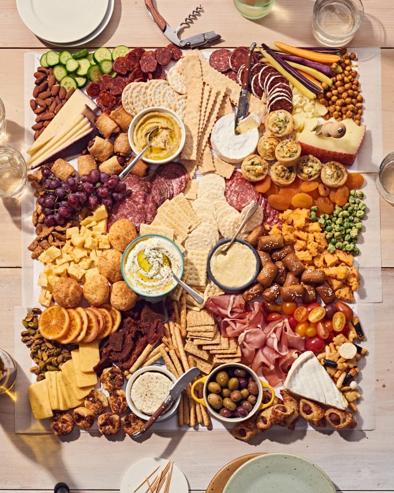 grazing table or board on a light wood table. There is meat, cheese, crackers, fruit, vegetables, dips, olives, and more. Plates and glasses, toothpicks on the table by the food.