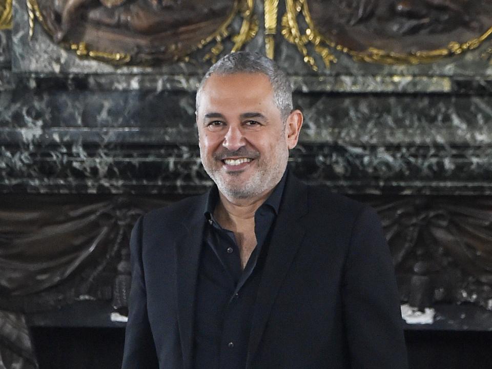 elie saab, a middle aged man with trimmed grey facial hair and short grey hair, smiling in front of a grey background. he's wearing an all black suit, with an open jacket and no tie.