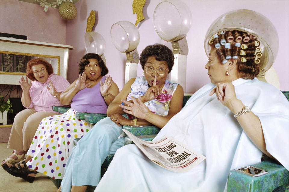Four women in a salon with one under a hairdryer, looking surprised, with a magazine visible