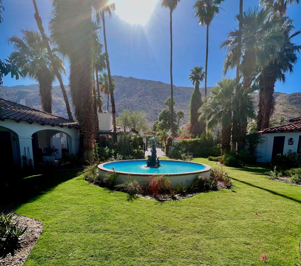 A fountain on a green lawn under palm trees next to mountains.