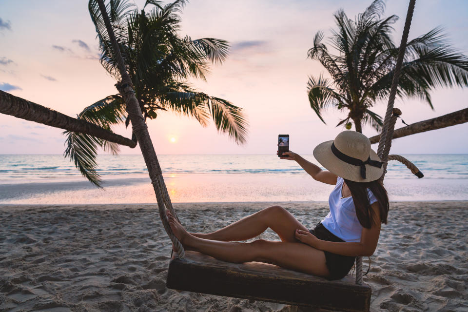 Summer travel vacation concept, Happy traveler asian woman with hat using mobile phone for selfie and relax on swing in tropical beach at sunset, Koh Kood, Thailand