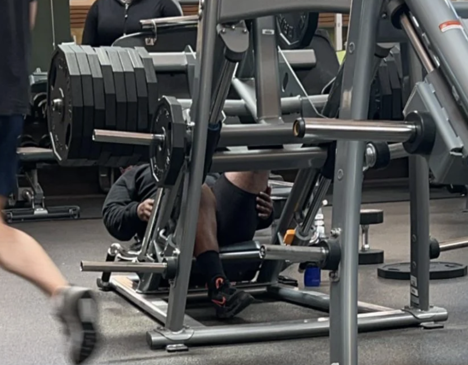 Person in gym using a leg press machine with several weight plates loaded. Another person walks by the equipment