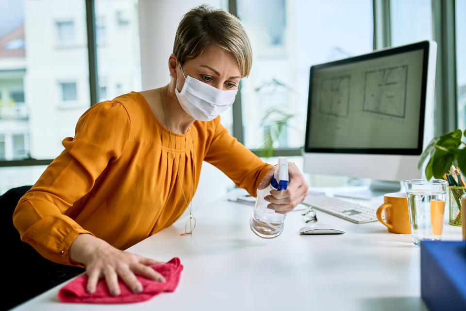 Businesswoman with face mask disinfecting her desk while working in the office during virus epidemic.