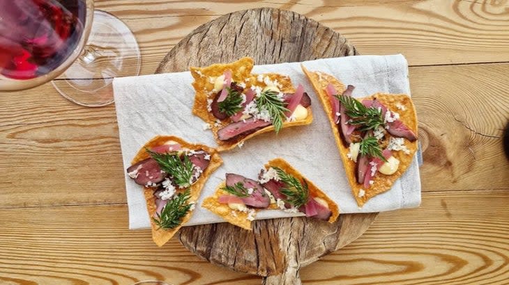 A circular wooden platter with crudites of grilled beef heart atop crackers of yellow peas, with a glass of red wine to the left