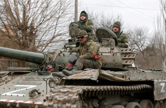 Three service members of pro-Russian troops in Ukraine in uniforms without insignia, one wearing red ribbons around his left arm and right leg, ride a tank marked