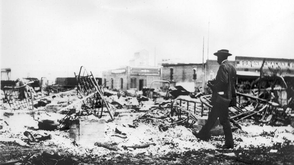 An African American man with a camera looking at mangled bed frames, which rise above the ashes of a burned-out block after the Tulsa Race Massacre, Tulsa, Oklahoma, June 1921. - Oklahoma Historical Society/Getty Images