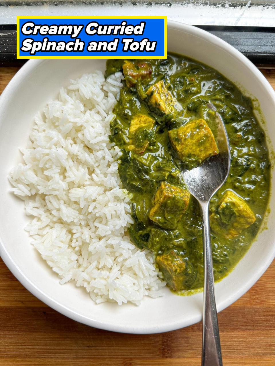 A white bowl with rice and palak paneer, an Indian dish with spinach and cheese cubes, placed on a wooden surface