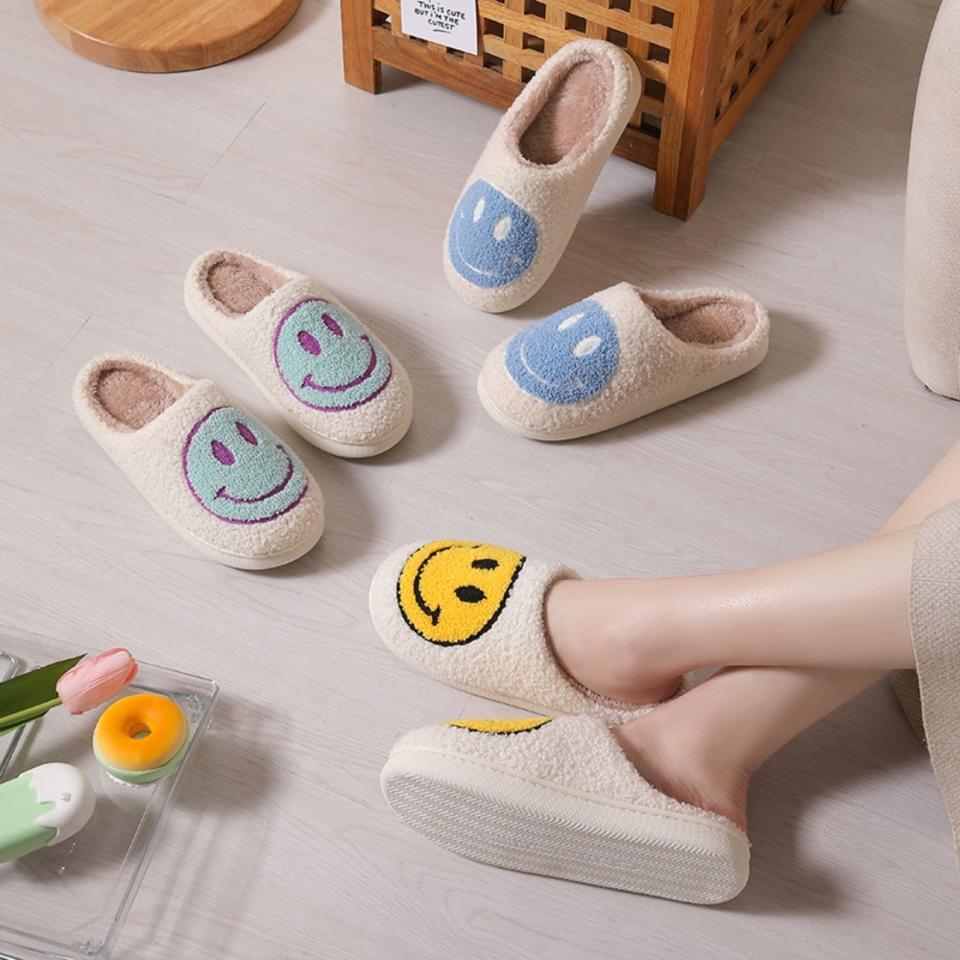 A person's feet wearing one smiley face slipper, with other slippers nearby on a floor