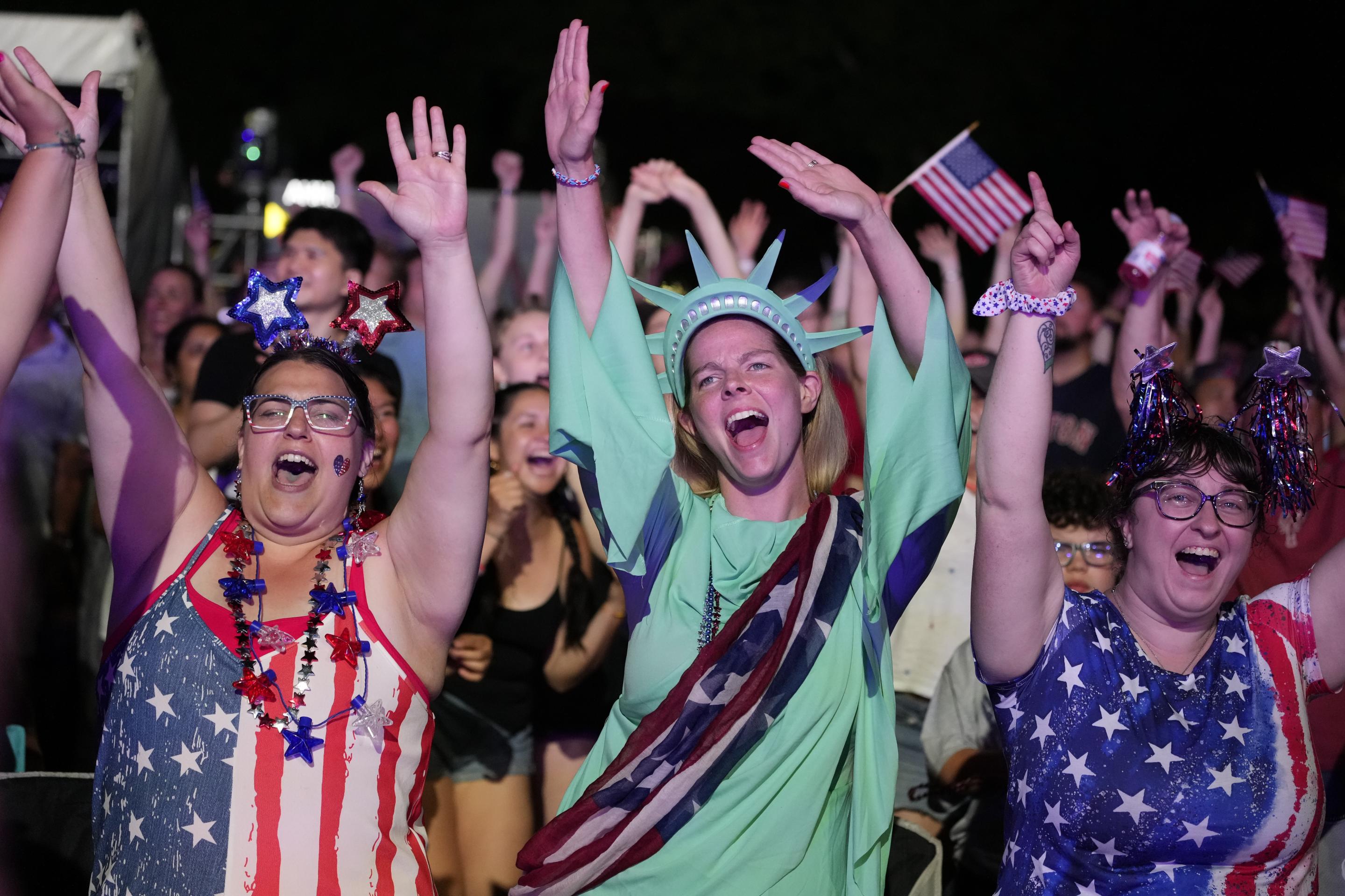 A woman in a Statue of Liberty costume standing alongside other women dressed in shirts with stars-and-stripes patterns raise their hands in the air and cheer during the Boston Pops Fireworks Spectacular in Boston.