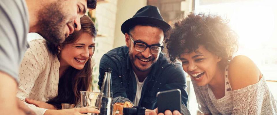 Portrait of cheerful young friends looking at smart phone while sitting in cafe. Mixed race people sitting at a table in restaurant using mobile phone.