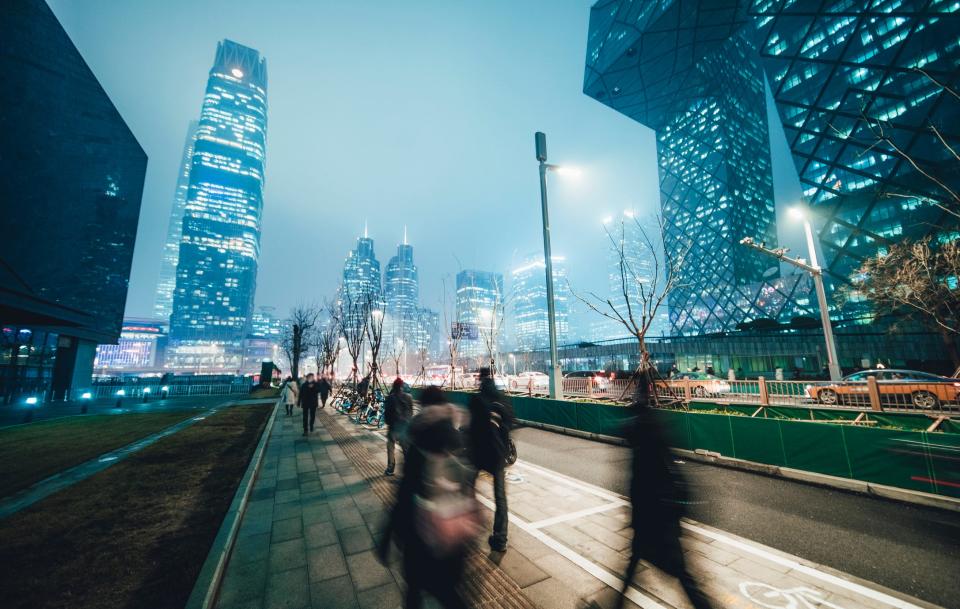 An evening cityscape in the background behind a small urban park in as blurred pedestrians are walking quickly along the sidewalk.