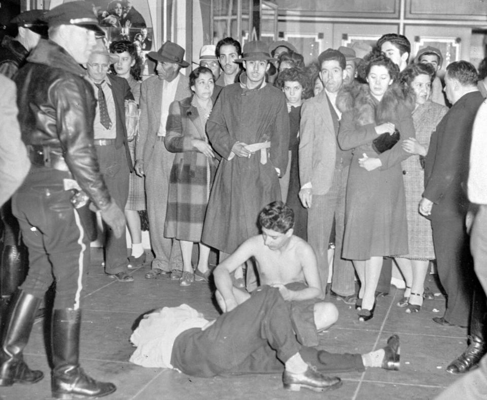 A woman kneels beside a man lying on the ground; a policeman looks on.