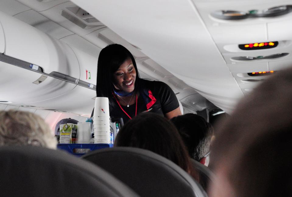 A flight attendants with a cart carrying cups and juice boxes smiles at an unseen passenger