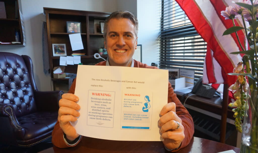 Rep. Andrew Gray, D-Anchorage, in his capitol office on May 16, 2024, holds up a printout showing how warning signs about alcohol consumption's health effects would change under legislation he sponsored. (Photo by Yereth Rosen/Alaska Beacon)