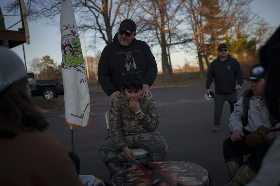 John Johnson, tribal president of the Lac du Flambeau Band of Lake Superior Chippewa Indians, stands behind grandson Ganebik Johnson as he prepares to drum and sing at a youth spearfishing event Saturday, April 20, 2024, in Lac Du Flambeau, Wis. (AP Photo/John Locher)