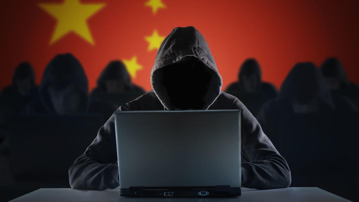  A group of 7 hackers, 6 slightly blurred in the background and one in the foreground, all wearing black with hoods pulled up over their heads. You cannot see their faces. The hacker in the foreground sits with an open laptop in front of them. The background, behind the hackers, is a Chinese flag. 