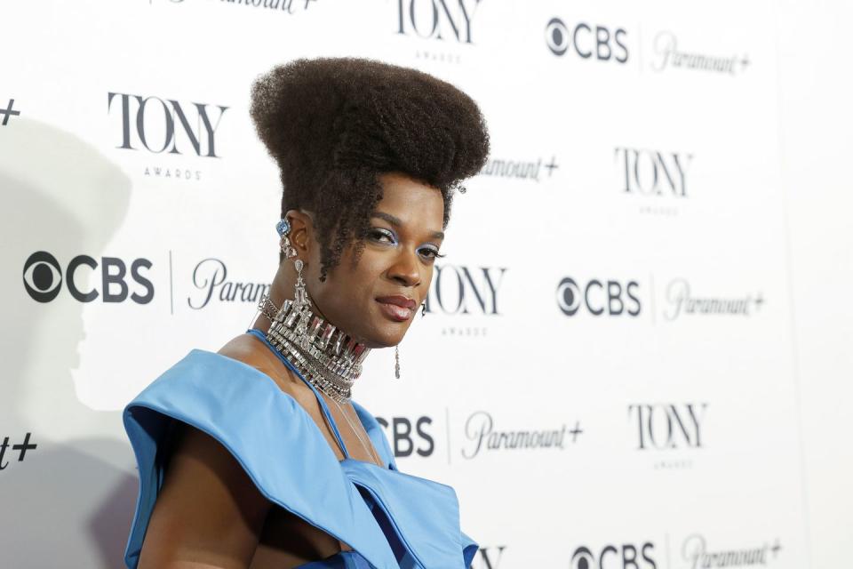 j harrison ghee, wearing a red dress, looks at the camera while posing in front of a white wall with tony awards and cbs logos on it