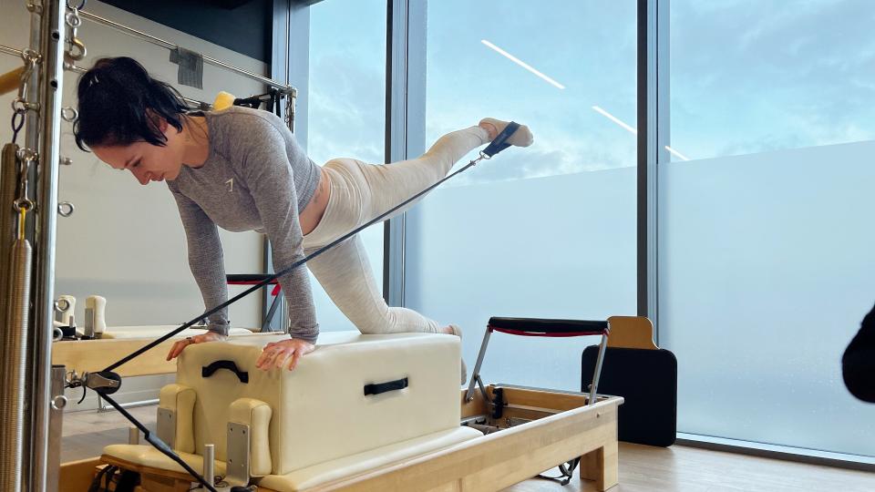 Writer Sam on a reformer Pilates bed on hands and one knee extending her left leg behind her, front view