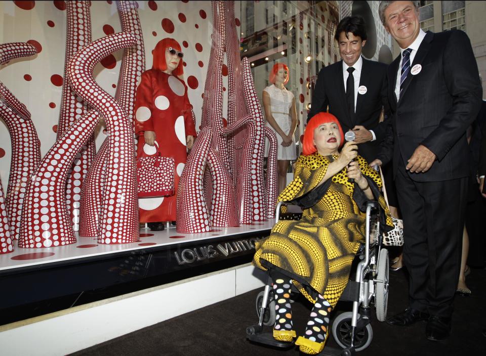 FILE - In this July 10, 2012 file photo, Japanese artist Yayoi Kusama in a red wig speaks with Louis Vuitton's second in command Jordi Konstans, left, and CEO Yves Carcelle, right, in front of the windows of Vuitton's flagship store for the unveiling of a new collaborative collection by Vuitton creative designer Marc Jacobs and Kusama in New York. Kusama's signature splash of dots has now arrived in the realm of fashion in a new collection from French luxury brand Louis Vuitton - bags, sunglasses, shoes and coats. The latest Kusama collection is showcased at its boutiques around the world, including New York, Paris, Tokyo and Singapore, sometimes with replica dolls of Kusama. (AP Photo/Kathy Willens, File)