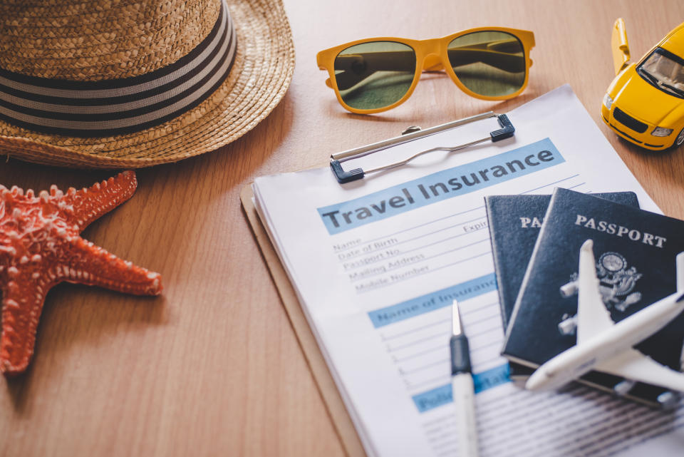 Travel essentials—straw hat, sunglasses, toy car, passport, toy plane, pen, starfish, and a clipboard with a travel insurance form—laid on a wooden surface
