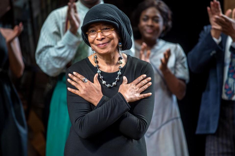 A woman stands onstage, her arms crossed over her chest, as people stand behind her applauding.