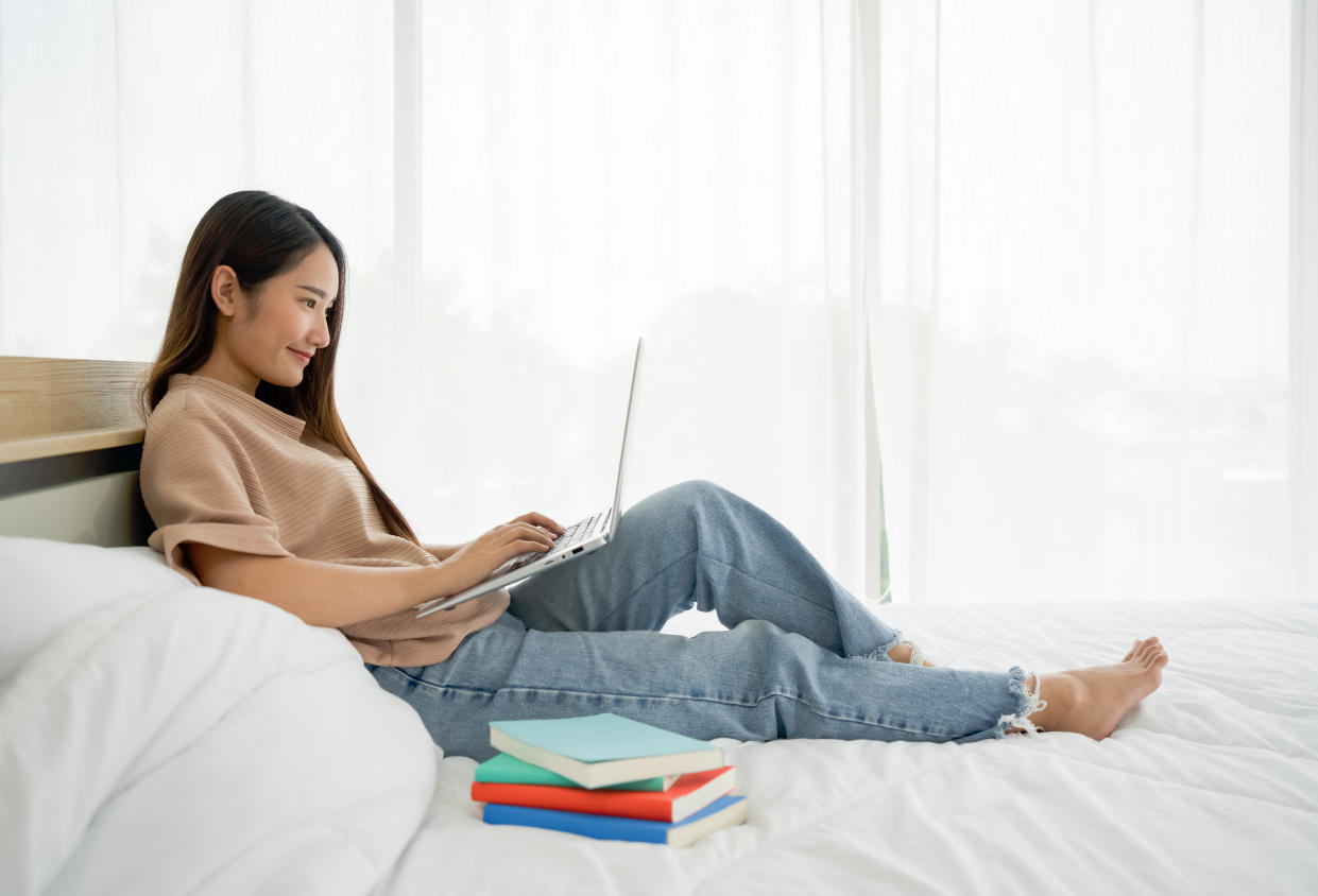 Young asian woman with a smile typing on laptop computer keyboard while relax on bed at home, after reviewing the textbook.