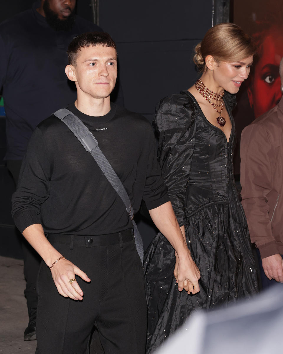 Tom Holland and Zendaya hold hands. Tom is wearing a casual outfit with a strap over his shoulder, while Zendaya is in a dramatic, voluminous dress with puffed sleeves