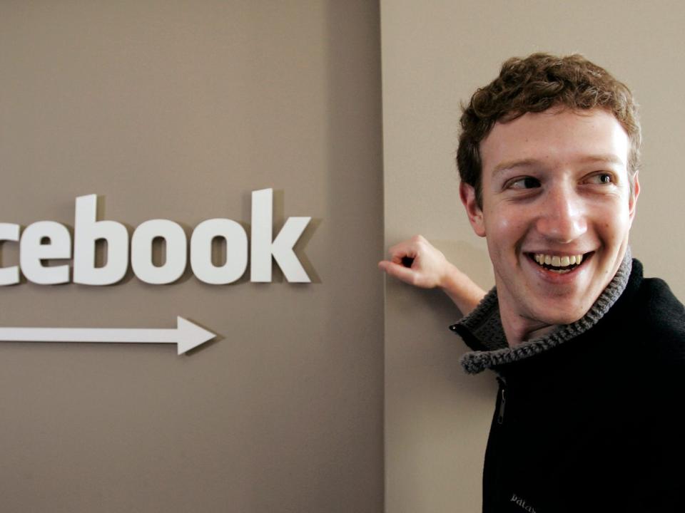 Mark Zuckerberg smiles while standing next to sign that says Facebook with arrow underneath