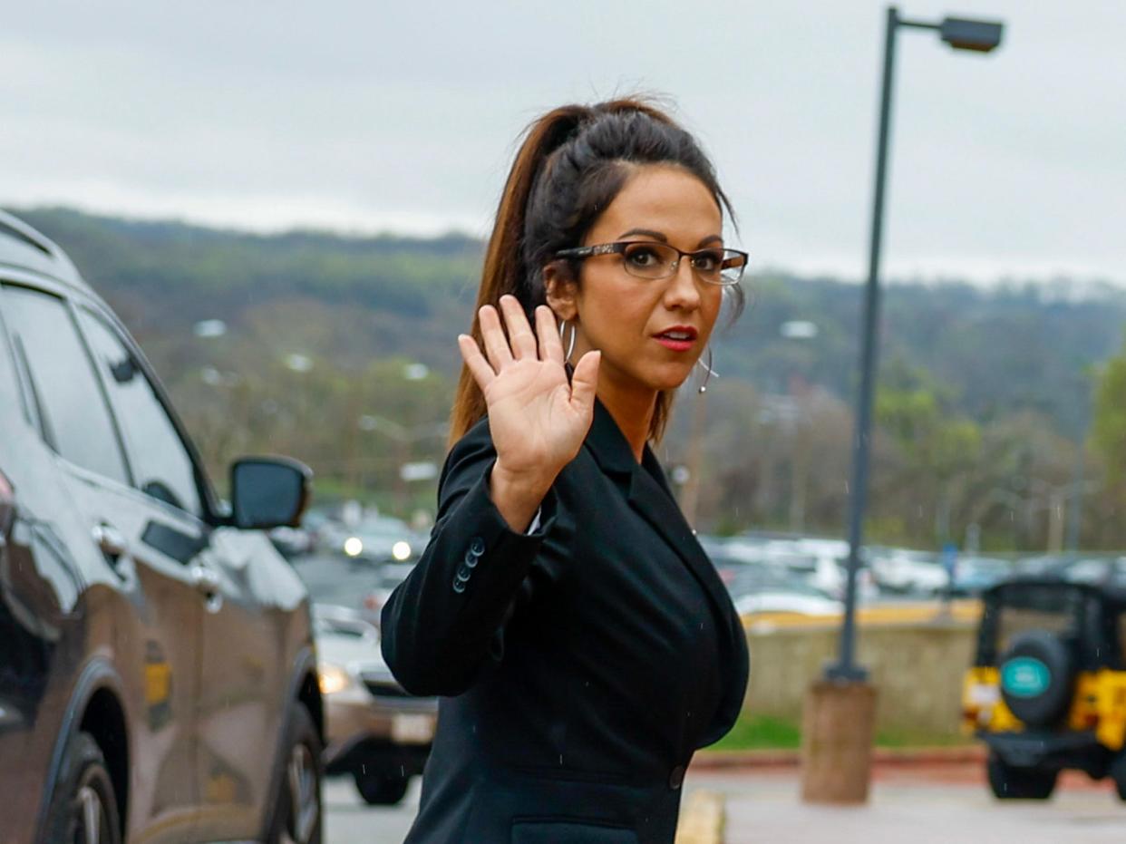 Rep. Lauren Boebert waving in a parking lot while standing next to a car and wearing a black suit and glasses.