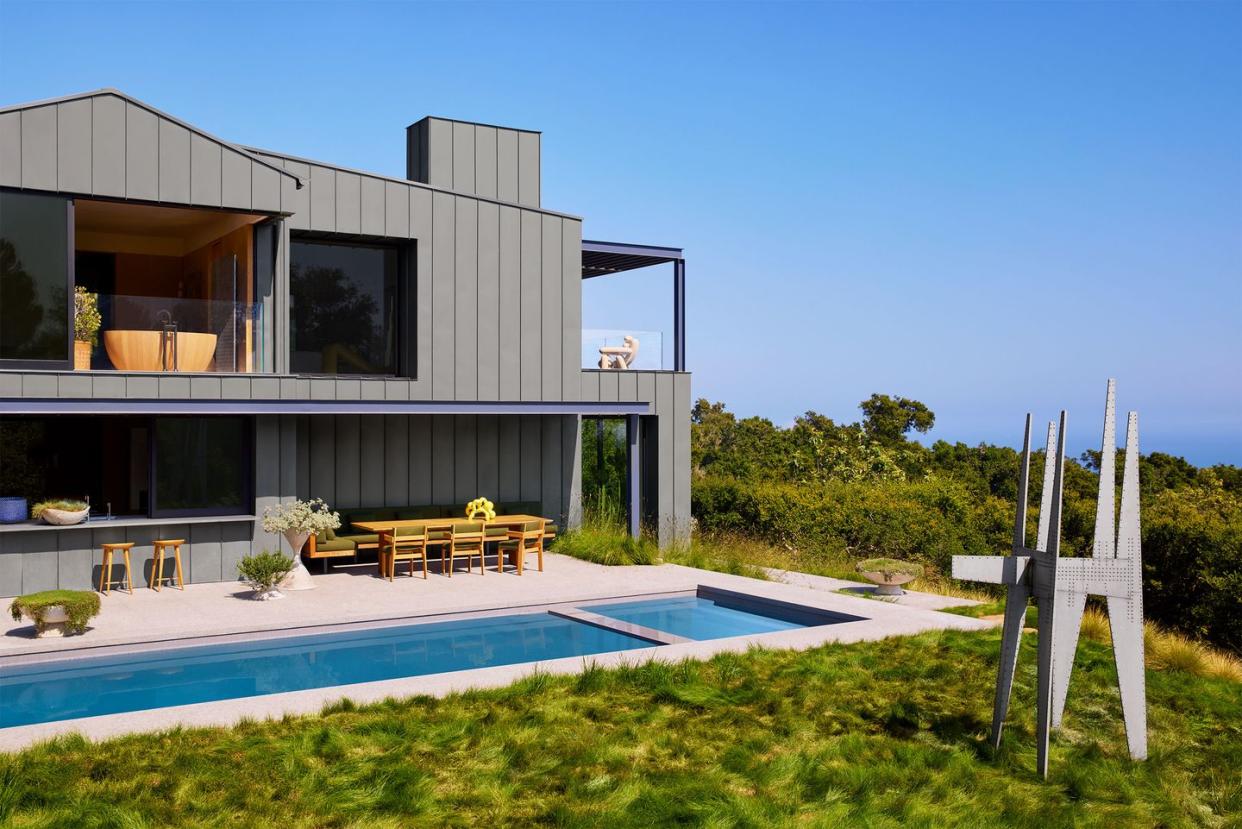 on the 2nd floor of the back of the house are two windows, one showing a bathtub, and a terrace, outside the 1st floor is a dining table with chairs next to a built in lap pool, and a metal sculpture