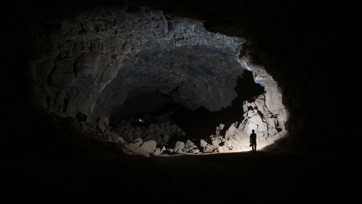  A large cave-like lava tube that is dark and underground. We see a researcher with a light in the corner. 