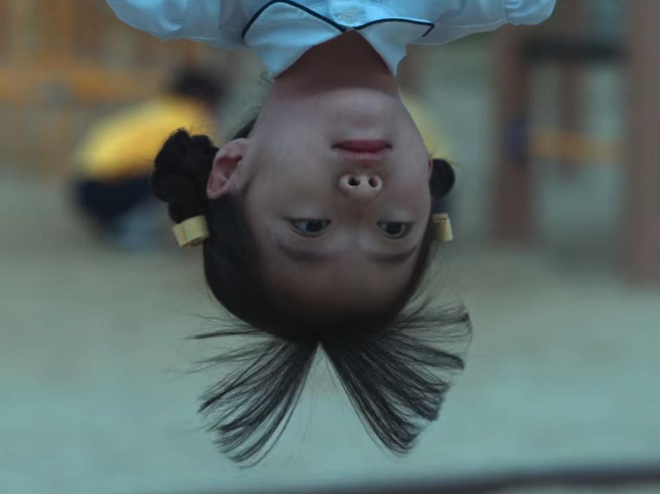 a young girl hanging upside down on a playground, her straight bands hanging down from her head. she has a neutral, curious expression on her face