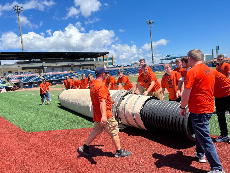 Members of the Blue Wahoos front office staff, wearing Zarzaur Tarp Crew T-shirts for the sponsor company, prepare to roll out the field tarp during a test run prior to 2023 season. The tarp will soon be donated in a contest.