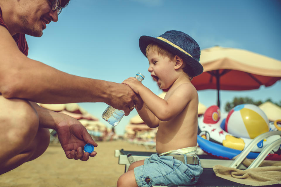 A man helps a young boy in a sun hat open a bottle of water at the beach. They are surrounded by beach umbrellas and toys