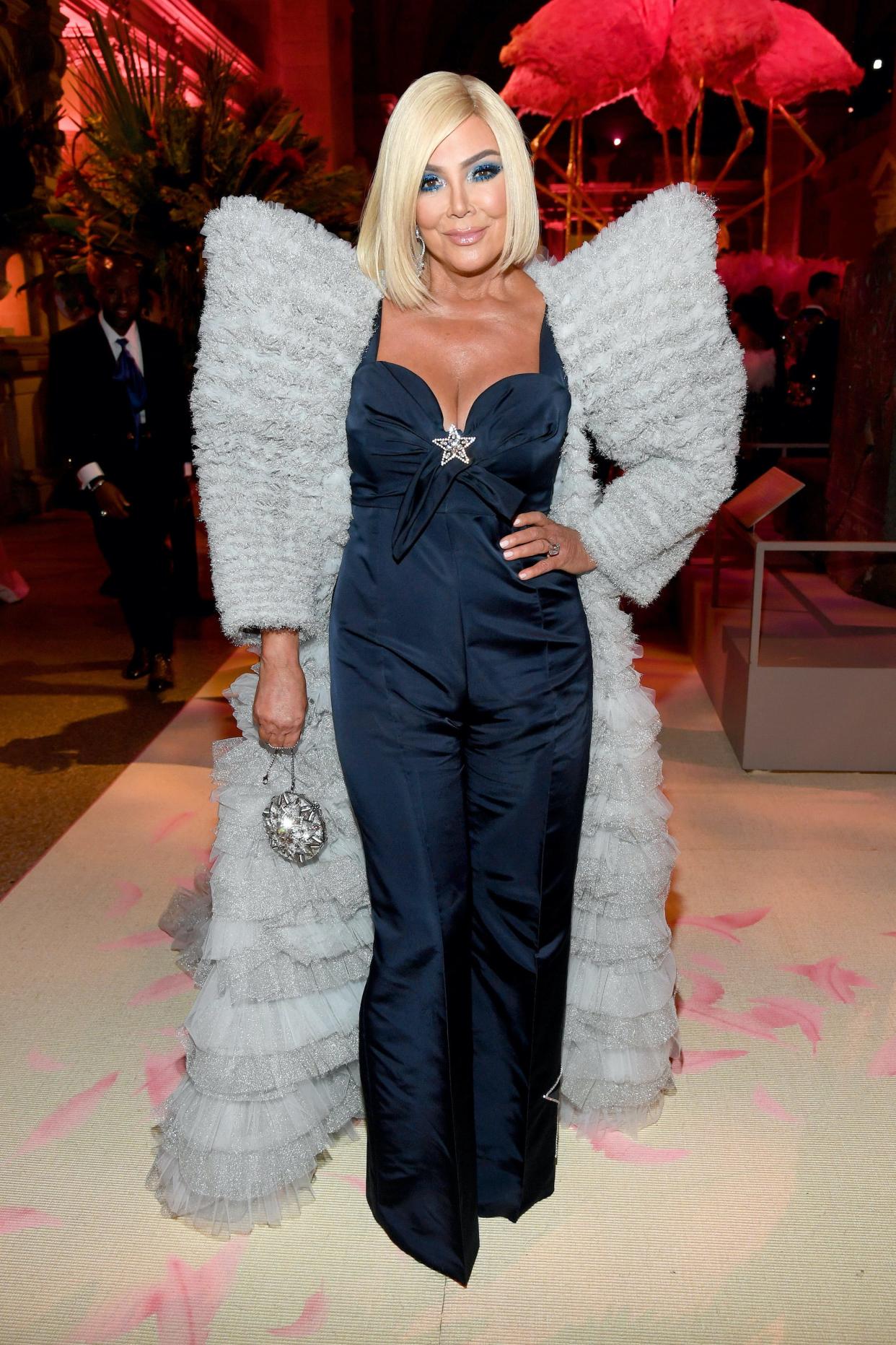 Kris Jenner attended the 2019 Met Gala in a blue jumpsuit with exaggerated shoulders and a silver star at her cleavage. Her hair is blonde and shoulder length.