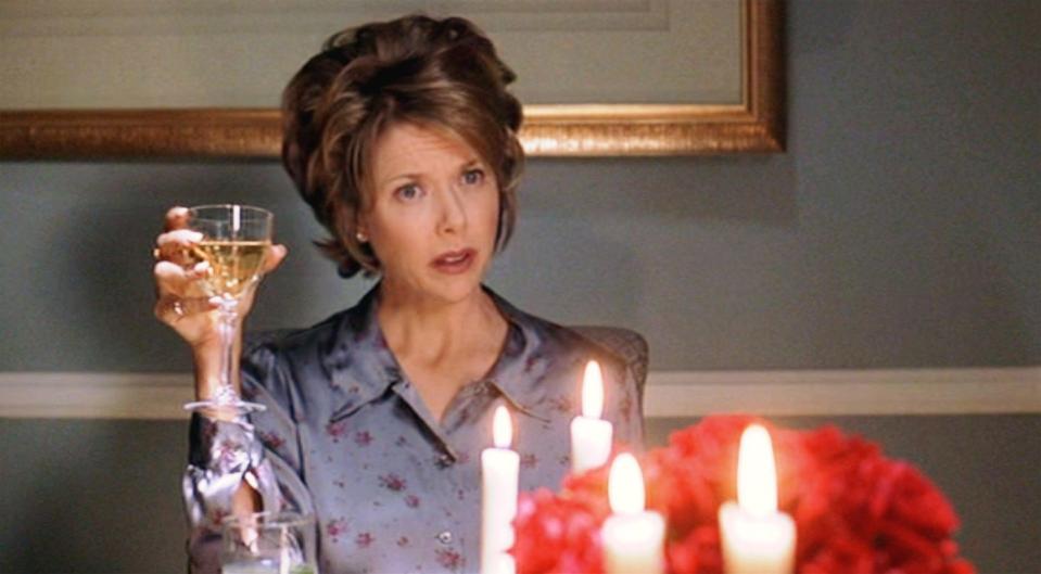 annette bening in character for american beauty, she sits at a dining table and holds a glass of wine up, in front of her are lit candles and red flowers